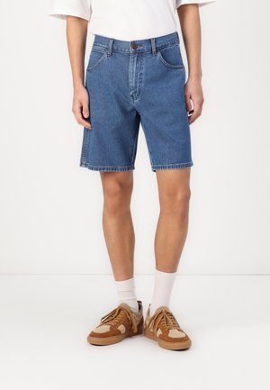 Denim shorts in medium blue with a straight cut, five pockets, and contrast stitching. Worn with brown sneakers and white socks.