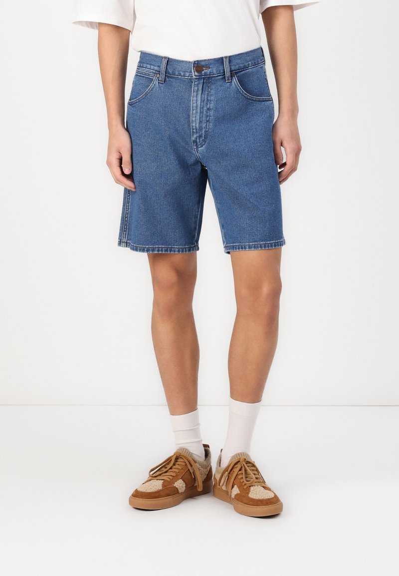 Shorts de mezclilla en azul medio con un corte recto, cinco bolsillos y costuras de contraste. Llevados con zapatillas marrones y calcetines blancos.