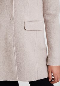 Cappotto di lana beige texturizzato con tasca a pattina, chiusure a bottone e maniche lunghe indossato sopra pantaloni neri. Mano con piccoli tatuaggi visibili.