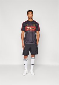 Camisa de fútbol negra con acentos rojos y logo, combinada con pantalones cortos negros, medias blancas con rayas negras y botas blancas, de pie.