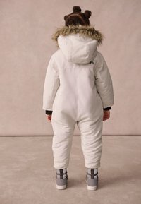 White padded snowsuit with a fur-lined hood, ribbed black cuffs, and gray boots. Features a rear logo detail and a smooth texture.