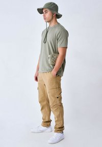 T-shirt verde oliva, pantaloni cargo beige con tasche laterali, sneakers bianche e un cappello a secchiello verde oliva abbinato. Borsa strutturata tenuta in mano.