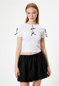 Weißes, kurzes T-Shirt mit gerippter Textur, das drei schwarze Schleifen und Aussparungen an den Schultern aufweist. Kombiniert mit einem schwarzen, gerafften Rock.