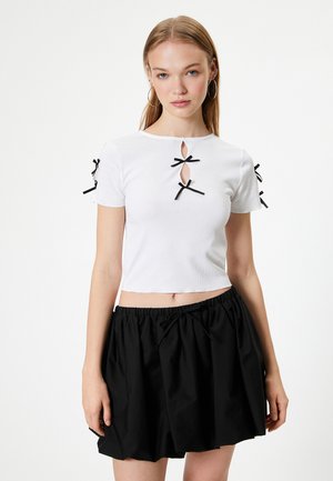 Weißes, kurzes T-Shirt mit gerippter Textur, das drei schwarze Schleifen und Aussparungen an den Schultern aufweist. Kombiniert mit einem schwarzen, gerafften Rock.
