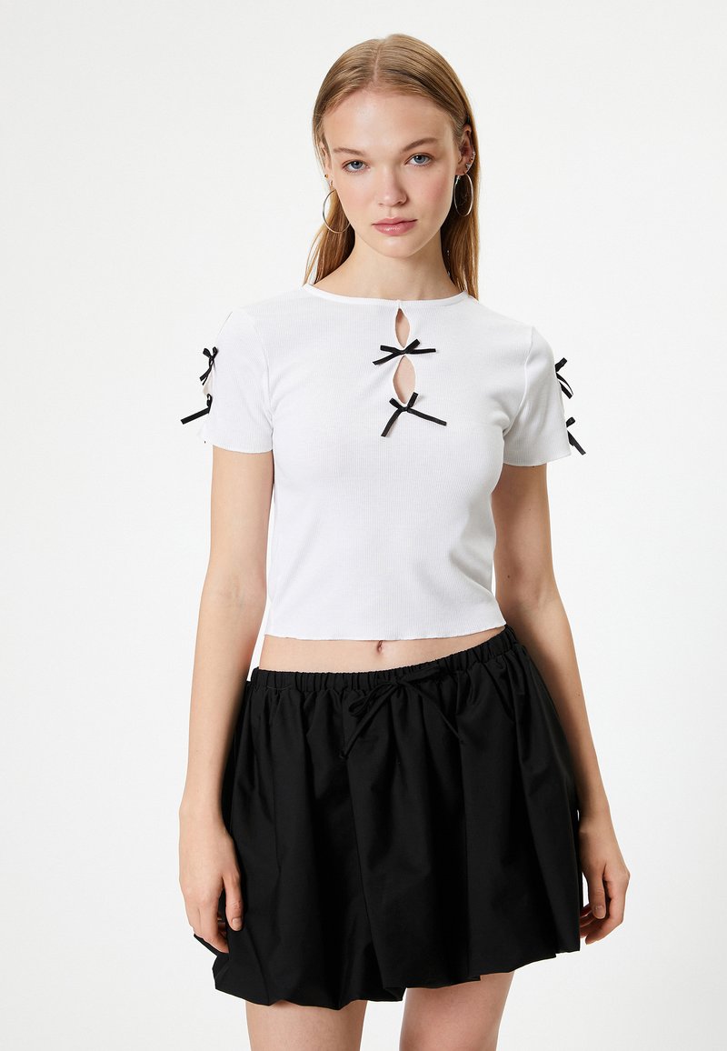 Weißes, kurzes T-Shirt mit gerippter Textur, das drei schwarze Schleifen und Aussparungen an den Schultern aufweist. Kombiniert mit einem schwarzen, gerafften Rock.