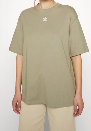 Personne portant un T-shirt Adidas ample de couleur vert olive avec un logo blanc centré sous l'encolure et un pantalon beige clair.