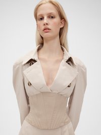 Beige trenchcoat med dyp V-hals, oversized krage og strukturert korsettlignende midje. Har store knapper og ribbede detaljer.