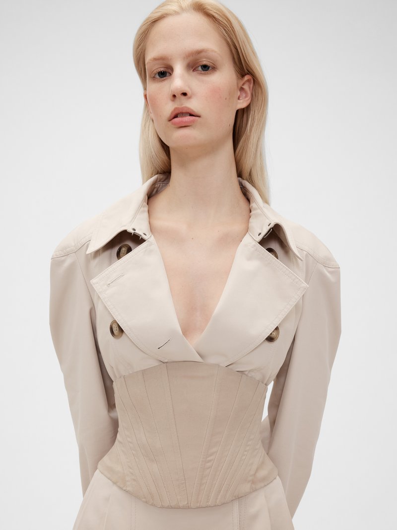 Beige trenchcoat med dyp V-hals, oversized krage og strukturert korsettlignende midje. Har store knapper og ribbede detaljer.
