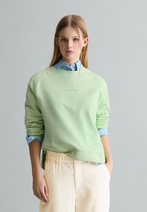 Młoda kobieta stoi ubrana w jasnooliwkową bluzę z kapturem na niebieskiej koszuli z kołnierzykiem oraz kremowe spodnie na tle jednolitego szarego tła.