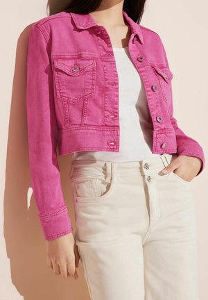 Femme portant une veste en jean courte rose vif sur un top blanc avec un pantalon taille haute écru, main dans la poche sur un fond pâle.