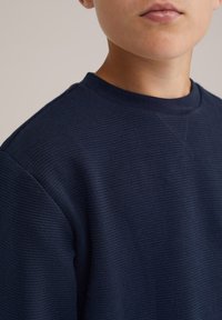 Navy mėlynas tekstūruotas džemperis su apvalia iškirpte ir gremžtiniu audiniu. Išskirtinis dėl subtilaus rašto ir tvarkingų siūlių detalių.