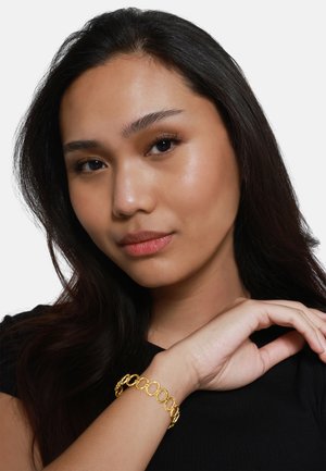 Eine Goldmetallarmband mit miteinander verbundenen ovalen Gliedern, am Handgelenk getragen. Einfarbig und mit glatter Textur, vor einem schwarzen Hemd und einem neutralen Hintergrund.