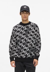 Melnā adīto džemperis ar baltu ģeometrisku logotipa rakstu, apaļa kakla izgriezums un ribota apkakle. Valkāts ar melniem džinsiem un saskaņotu cepuri.