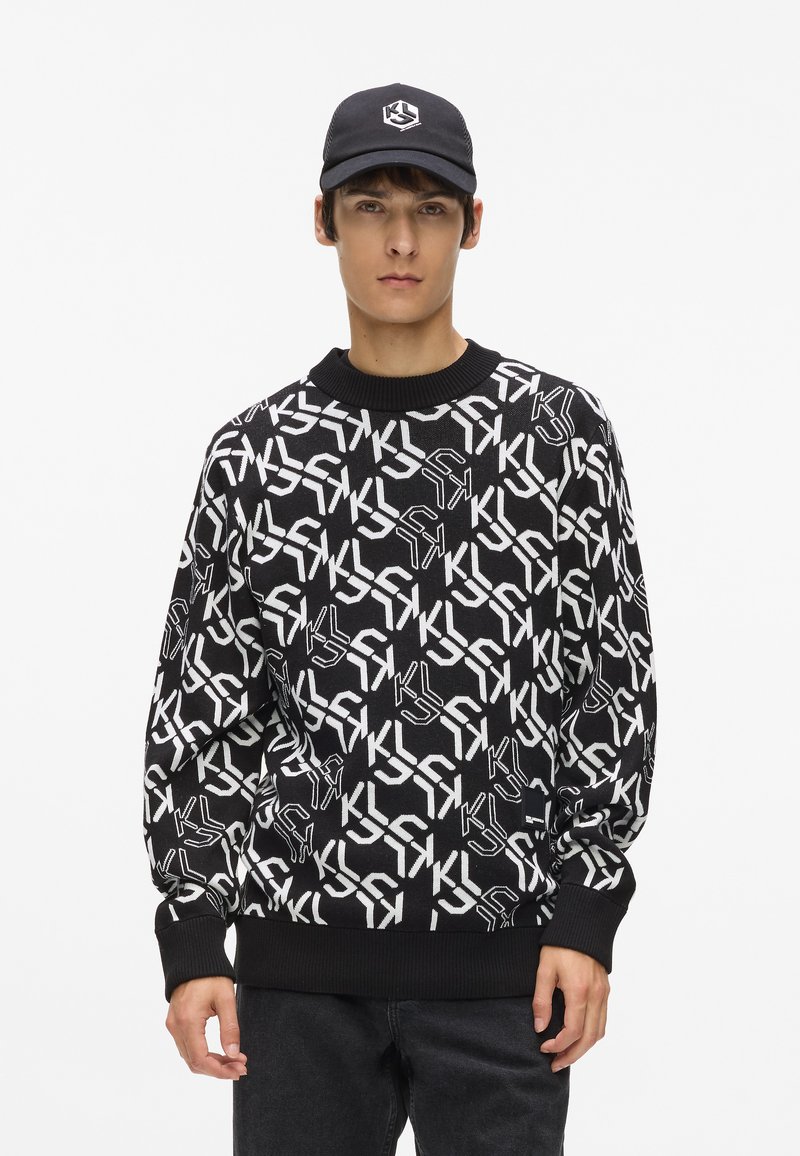 Melnā adīto džemperis ar baltu ģeometrisku logotipa rakstu, apaļa kakla izgriezums un ribota apkakle. Valkāts ar melniem džinsiem un saskaņotu cepuri.