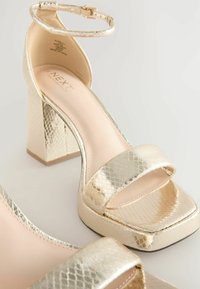 Arany színű, blokksarkú szandálok texturált, kígyóbőr hatású felülettel. Széles pánt a lábujjak felett és egy bokapánt jellemzi. Belső felületük sima.