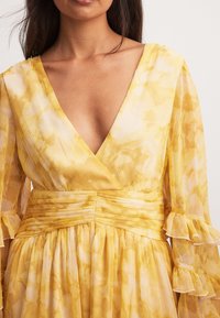 Keltainen kukkamekko, jossa on syvä V-pääntie, pleatattu vyötärö ja röyhelöiset läpikuultavat hihat. Kangas on kevyt ja ilmava tekstuuri ja vesivärikuvio.