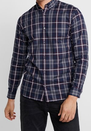 Uomo che indossa una camicia a quadri blu navy a maniche lunghe e pantaloni neri, in piedi con le braccia rilassate lungo i fianchi.