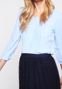 Blusa azzurro chiaro con maniche 3/4, caratterizzata da pieghe verticali sulla parte anteriore. Abbinata a una gonna scura e strutturata con un effetto a strati.