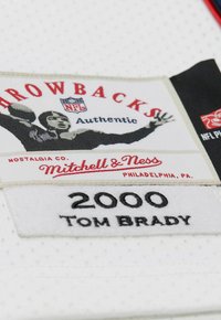 Weißes Stoffetikett mit NFL-Logo, rotem und schwarzem Text: "Throwbacks", "Authentic", "Mitchell & Ness, Philadelphia, PA", "2000 Tom Brady."