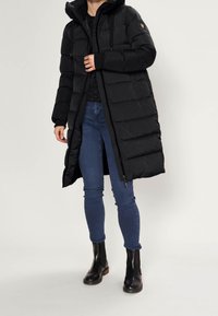 Kvinna i en lång svart vadderad jacka med dragkedja, mörk tröja, blå tight jeans och svarta ankelstövlar, stående mot en enkel bakgrund.