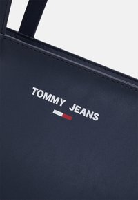 Tummansininen nahkainen olkalaukku, jossa valkoinen "TOMMY JEANS" -logo punavalkoisilla korostevyöhykkeillä. Sileä pinta ja tukevat olkahihnat.