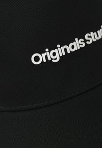 Mustmust koos struktureeritud disainiga, millel on tekstureeritud kangas ja silmapaistev valge "Originals Studio" logo ees.