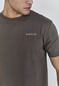 Brūna īsām piedurknēm T-krekls, izgatavots no gludas auduma, ar baltu "SIKSILK" logo uz krūšu daļas. Apaļa apkakle un vienkāršs dizains.