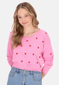Hellrosa Pullover mit V-Ausschnitt, dekoriert mit verstreuten roten Herzmustern. Weicher Stoff, lange Ärmel und ein cropped Design. Kombiniert mit leichter Jeans.