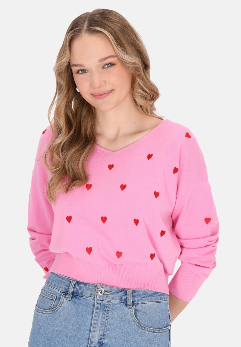 Hellrosa Pullover mit V-Ausschnitt, dekoriert mit verstreuten roten Herzmustern. Weicher Stoff, lange Ärmel und ein cropped Design. Kombiniert mit leichter Jeans.