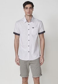 Camisa de botones de manga corta en gris claro con puños oscuros y un bolsillo en el pecho. Lleva puestos unos pantalones cortos de color beige arremangados. Tejido sutilmente texturizado.