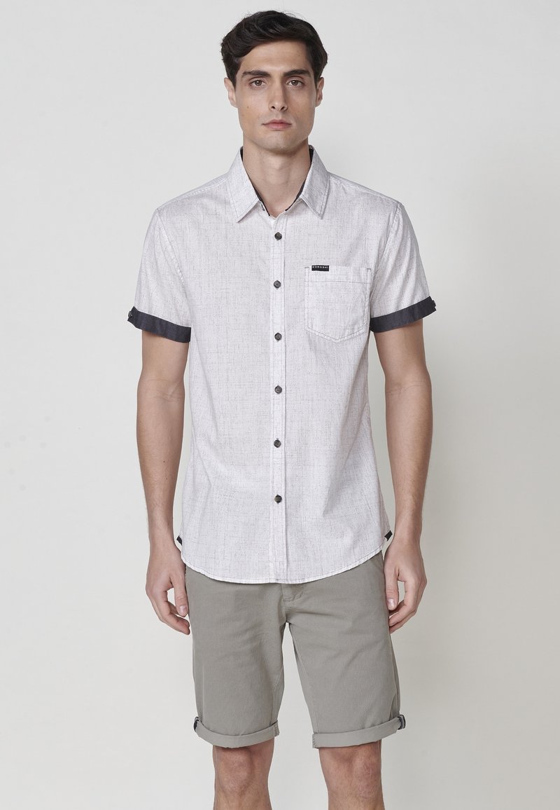 Camisa de botones de manga corta en gris claro con puños oscuros y un bolsillo en el pecho. Lleva puestos unos pantalones cortos de color beige arremangados. Tejido sutilmente texturizado.