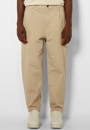Mann trägt beige zulaufende Hose und cremefarbene Sneaker, steht vor einem schlichten hellen Hintergrund.