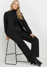 Svart strukturerad jacka över en svart polotröja, kombinerad med vida svarta byxor och ankelskor, sittande på en svart metallpall.