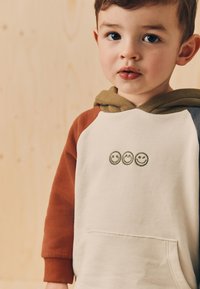 Mikina s kapucňou vyrobená z bavlny, s telom v krémovej farbe, hnedými rukávmi a tromi zelenými vyšívanými smajlíkmi na prednej strane.