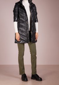 Kvinna i svart quiltad kappa, vit polotröja, olivgröna byxor och svarta klumpiga snörskor som står på ett beige golv.