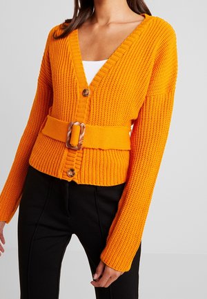 Femme portant un cardigan en tricot orange vif avec une grande boucle de ceinture par-dessus un haut blanc et un pantalon noir, vue des épaules aux hanches.