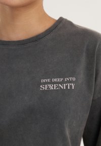 Dunkelgrauer Pullover mit rundem Ausschnitt und langen Ärmeln, auf dessen Vorderseite in weißer Schrift "DIVE DEEP INTO SERENITY" steht.