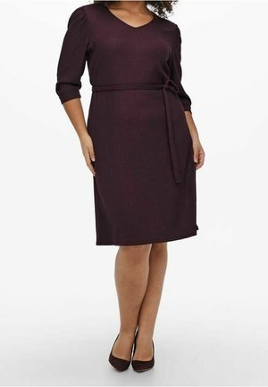 Femme debout portant une robe prune foncé longueur genou avec manches trois-quarts, col en V et ceinture nouée à la taille, associée à des chaussures assorties.