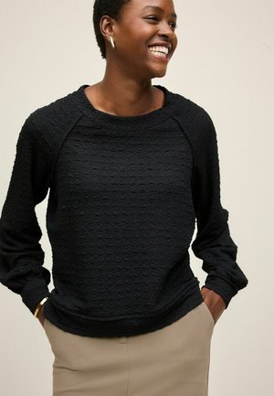 Black textured sweater with long sleeves and a round neckline, paired with beige pants. Features a subtle raised pattern and relaxed fit.