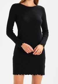 Black long-sleeve dress made of knit fabric, featuring a rounded neckline and a scalloped hem. Smooth texture, fitted silhouette.