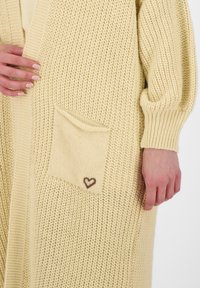 Sweter rozpinany w miękkim żółtym kolorze z teksturowanym wzorem, posiadający kieszeń ozdobioną małym emblematem w kształcie serca, zaprojektowany w luźnym fasonie.