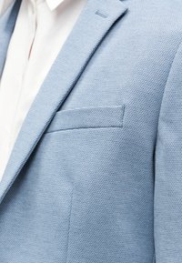 Φωτεινό μπλε σακάκι με υφή φορεμένο πάνω από λευκό πουκάμισο με γιακά, εμφανίζοντας τη θήκη στο στήθος και τις λεπτομέρειες στο πέτο.