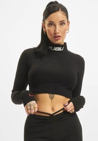 Čierny skrátený top s dlhým rukávom a vysokým golierom, ktorý má biele logo a bezprstové manžety. Model nosí zodpovedajúce čierne nohavice.