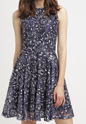 Robe bleu marine sans manches avec un motif de dentelle florale blanche et une taille ajustée, portée par une personne aux cheveux bruns mi-longs.