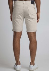 Lys beige shorts med rullekanter, laget av bomull. Har to baklommer og et logo-patch på livlinjen. Strukturerstoff.