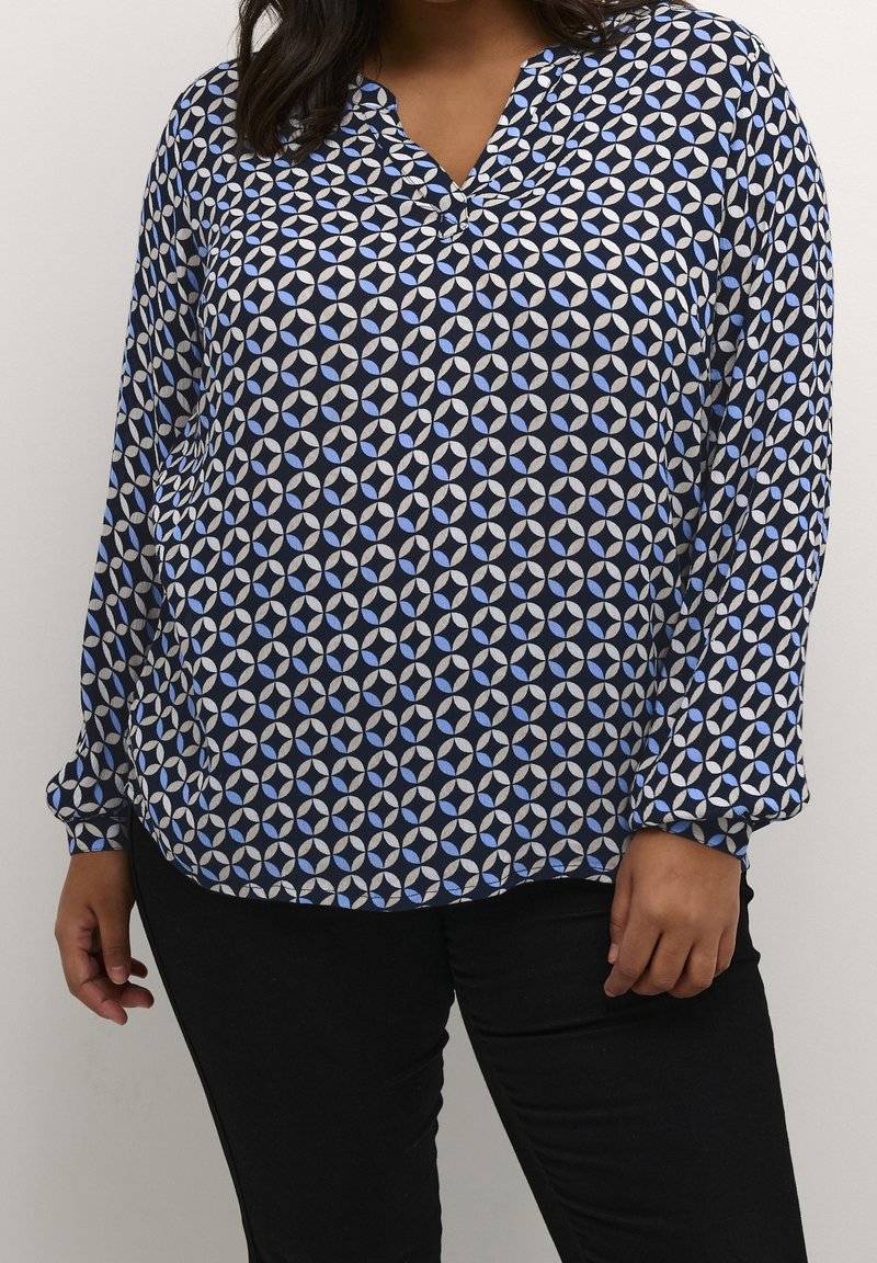 Femme portant un chemisier à manches longues avec un motif géométrique circulaire bleu, blanc et noir et un pantalon noir sur un fond uni.