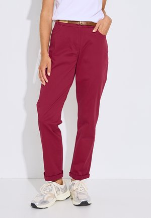 Persona con pantalones rojo oscuro remangados, cinturón marrón, camiseta blanca y zapatillas blancas y beige, de pie frente a un fondo sencillo.