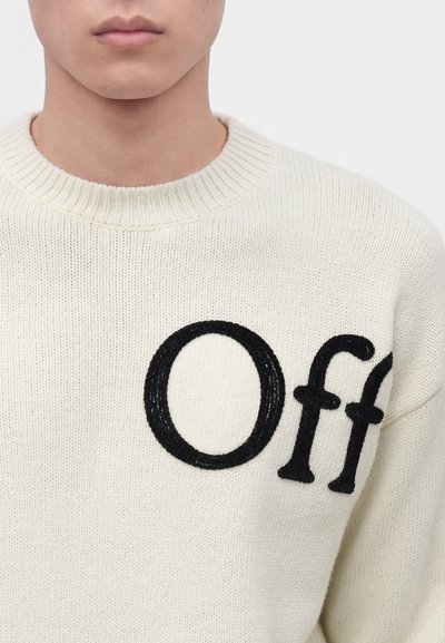 Bas du visage et haut du torse d'une personne portant un pull en maille blanche avec des lettres noires "Off" sur la poitrine.