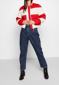 Giacca bomber a righe rossa e crema con chiusura a zip, colletto a costine e orlo elasticizzato, abbinata a jeans blu scuro a vita alta e stivali neri.