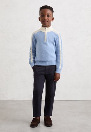 Dreng står med hænderne i lommerne iført en lyseblå og cremefarvet kvartlynlås sweater, mørke bukser og brune sko mod en ensfarvet baggrund.